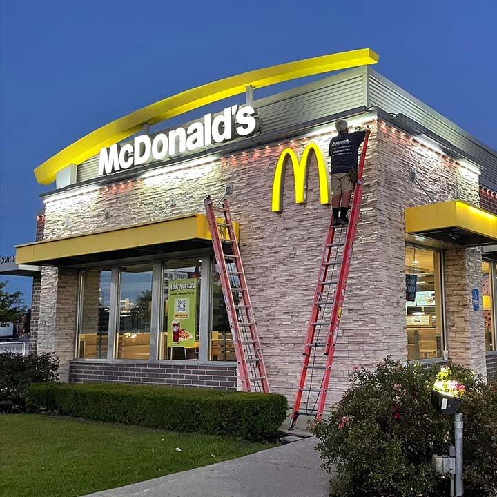East-Bay-Trimlight-comercial lighting A worker on a ladder repairs the lighting on a McDonalds sign at dusk, possibly performing a permanent installation, with another ladder leaning against the building and bushes in the foreground.
