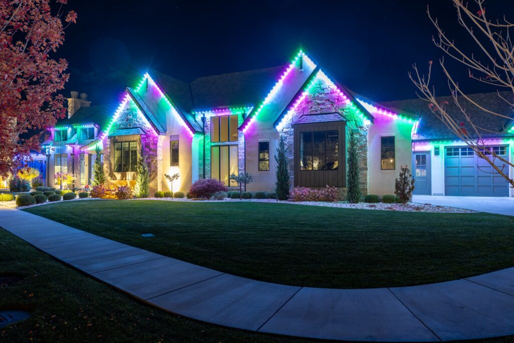 A house at night decorated with bright, colorful LED lights along the rooflines showcases a stunning permanent lighting installation, illuminating the exterior in green, blue, and purple hues. The lawn and landscaping are neatly maintained.