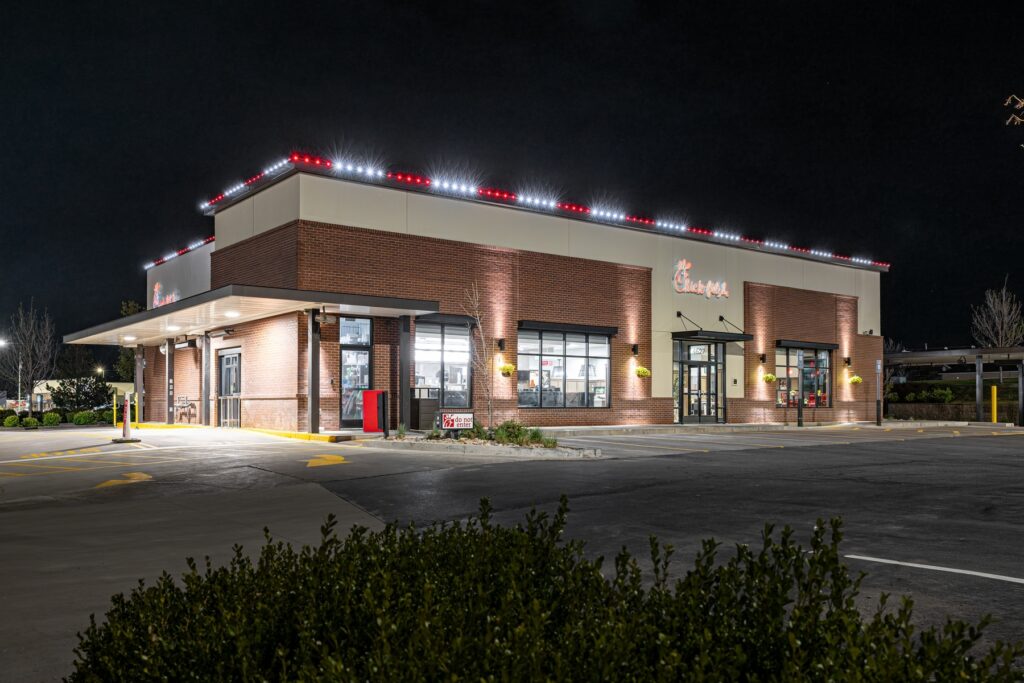A Chick-fil-A restaurant building is illuminated at night, with bright exterior house lights creating a festive atmosphere and a mostly empty parking lot in front. Some bushes are visible in the foreground, reminiscent of a holiday installation.
