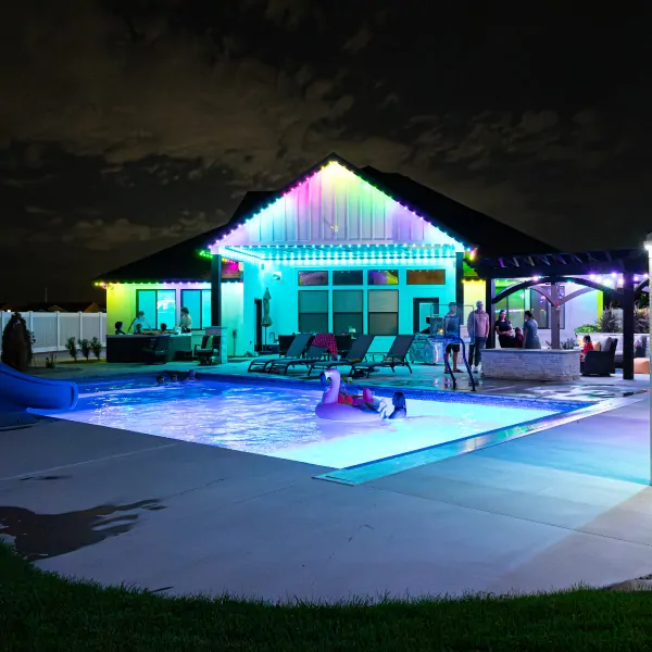 Attached image file A backyard pool at night is lit with colorful lights. People stand and swim, some using pool floats. The house in the background is decorated with permanent holiday house lights, creating a festive atmosphere.