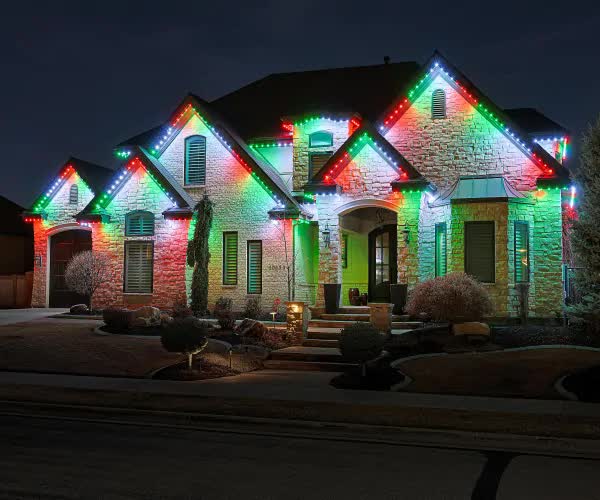 Attached image file A large house at night is decorated with red, green, and white holiday lighting along the roofline, illuminating the stone exterior and front yard landscaping.