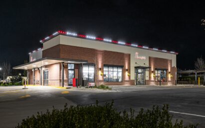 A Chick-fil-A restaurant building is illuminated at night, with bright exterior house lights creating a festive atmosphere and a mostly empty parking lot in front. Some bushes are visible in the foreground, reminiscent of a holiday installation.