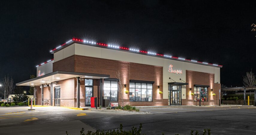 Attached image file A Chick-fil-A restaurant building is illuminated at night, with bright exterior house lights creating a festive atmosphere and a mostly empty parking lot in front. Some bushes are visible in the foreground, reminiscent of a holiday installation.