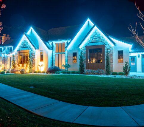 251020 151 W 710 S MAPLETON UT-25 (2) A large house at night is outlined with bright blue holiday lights along the roofline, featuring a permanent trimlight installation. The lawn is well-manicured, and the house is lit with warm exterior lights, creating a festive atmosphere.