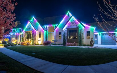 A house at night decorated with bright, colorful LED lights along the rooflines showcases a stunning permanent lighting installation, illuminating the exterior in green, blue, and purple hues. The lawn and landscaping are neatly maintained.