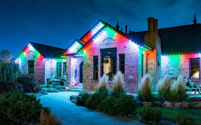 A brick house at night is decorated with colorful Christmas lights along the roofline, illuminating the walkway and garden plants in front of the home. The festive atmosphere is enhanced by a professional trimlight installation, ensuring the house lights shine beautifully throughout the holiday season.