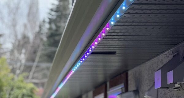 Attached image file A close-up view of a house gutter lined with multicolored LED house lights glowing in blue and purple, under a cloudy sky, with trees and a fence in the background—a festive installation perfect for the holiday season.